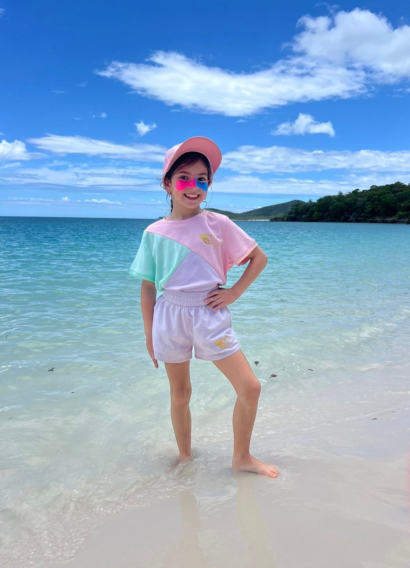 Girl wearing Dani Soccer Shirt in pink, green, and purple with Hero Athletica gold logo, standing on a beach.